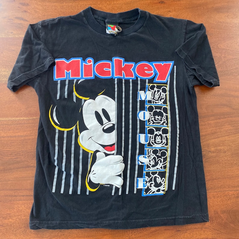 K&ouml;nnte beinhalten: Schwarzes T-Shirt mit einem Motiv von Mickey Mouse, der hinter einer Wand aus vertikalen Streifen hervorlugt. Der Text "Mickey Mouse" ist in roten und blauen Buchstaben &uuml;ber dem Motiv gedruckt.