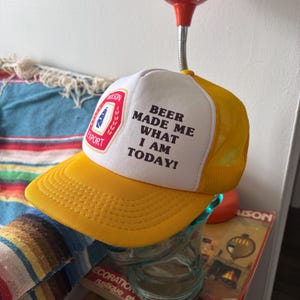 May include: Yellow and white trucker hat with the text "BEER MADE ME WHAT I AM TODAY!". The hat features a yellow brim and mesh back, with a patch that says "Wilson Export".