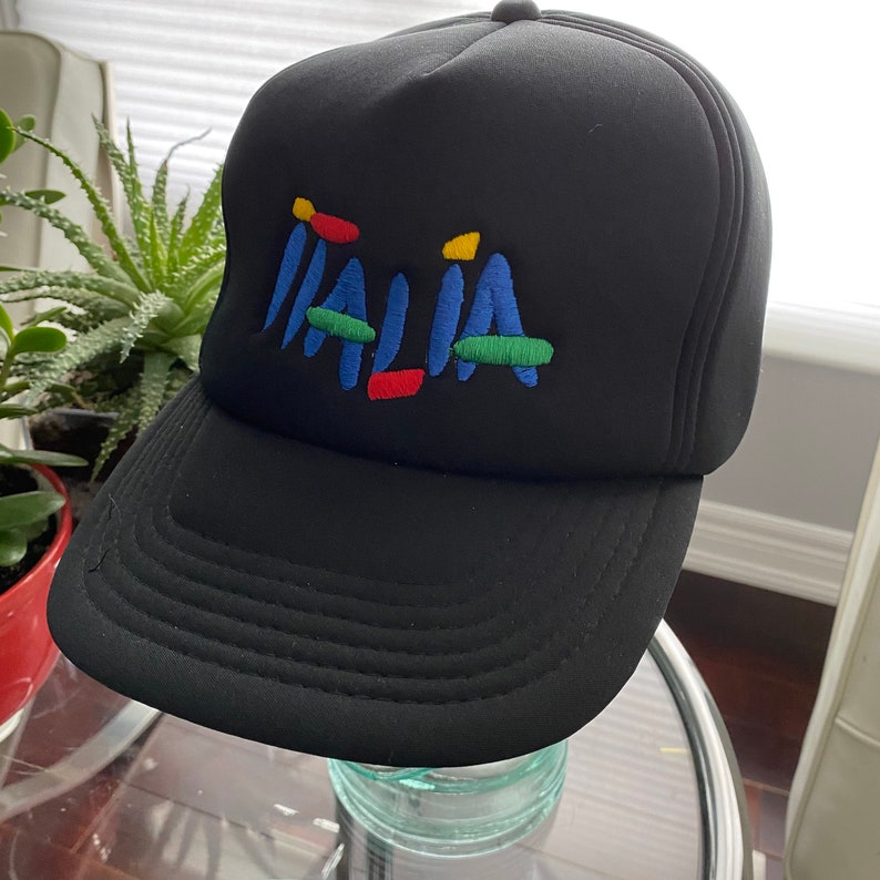 K&ouml;nnte beinhalten: Eine schwarze Trucker-Kappe mit einem bunten gestickten Logo, auf dem "ITALIA" steht.