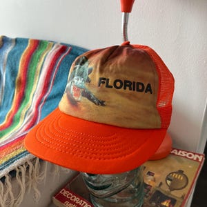 May include: Orange and brown mesh trucker hat with an alligator graphic and the word "FLORIDA" printed on the front.