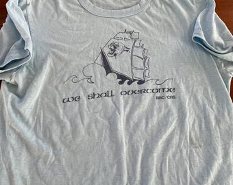 May include: Pale blue t-shirt with a navy blue graphic of a ship on waves. The words "we shall overcome" are printed below the ship, with "BBC/CHS" underneath. The shirt has a round neck and short sleeves.