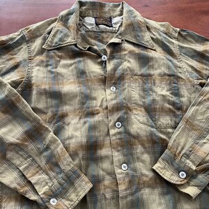 May include: A button-down shirt in a plaid pattern of olive green, brown, and blue. The shirt has a collar, a chest pocket, and white buttons. The fabric appears to be soft and worn.
