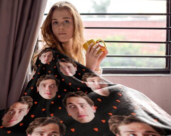 May include: A black fleece blanket featuring a repeating pattern of small red hearts and a repeated photographic portrait of a young man with brown hair. The blanket is draped over a surface, showcasing the custom printed design clearly.