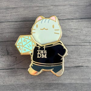 May include: A gold-colored enamel pin featuring a cat wearing a black hoodie with "THE DM" printed on it. The cat holds a teal D20 die. The pin has a light blue face and ears with gold accents. Several purple and blue dice are in the background.