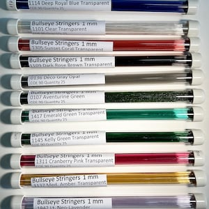 May include: A collection of Bullseye Stringers, 1 mm glass rods, in various colors including deep royal blue, clear, sunset coral, dark rose brown, and emerald green. Each tube is labeled with the color and product information.