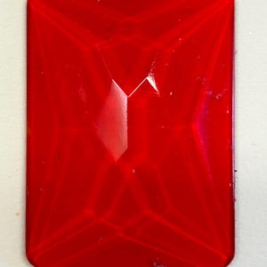 May include: A rectangular, faceted, red gemstone with rounded corners. The gem has a glossy finish and a central, triangular facet. The overall shape is reminiscent of a large, polished ruby.