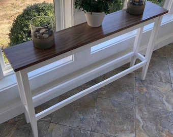 May include: A white console table with a wood top. The table has two shelves and is decorated with two glass jars filled with small stones. A potted plant sits on the top shelf.