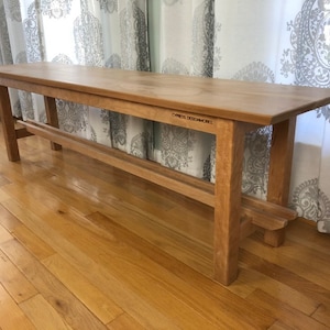 May include: A long, wooden bench with a simple design. The bench is made of light-colored wood and has a natural finish. The bench has a single, horizontal support beam underneath the seat. The bench is labeled "CYPRESS DESIGN & CABINETS".