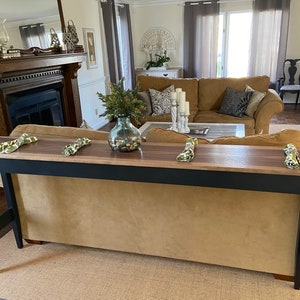 May include: A black and walnut console table with a black painted base and a walnut top. The table is placed in front of a brown sofa and has three green napkins on the top.