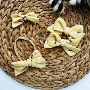 May include: A collection of yellow fabric hair bows with a black bee pattern. One bow is attached to a headband. The bows are arranged on a woven, circular placemat, with cotton stems in the background.