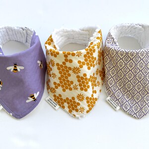 May include: Three baby bibs with different patterns. One is lavender with bee graphics, another is cream with a honeycomb design, and the third is gray with a geometric pattern. Each bib has a white, textured lining and a small tag.