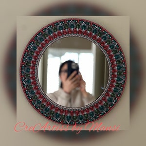 May include: A round mirror with a hand-painted mandala design in shades of red, green, and white. The mirror is surrounded by a black frame.