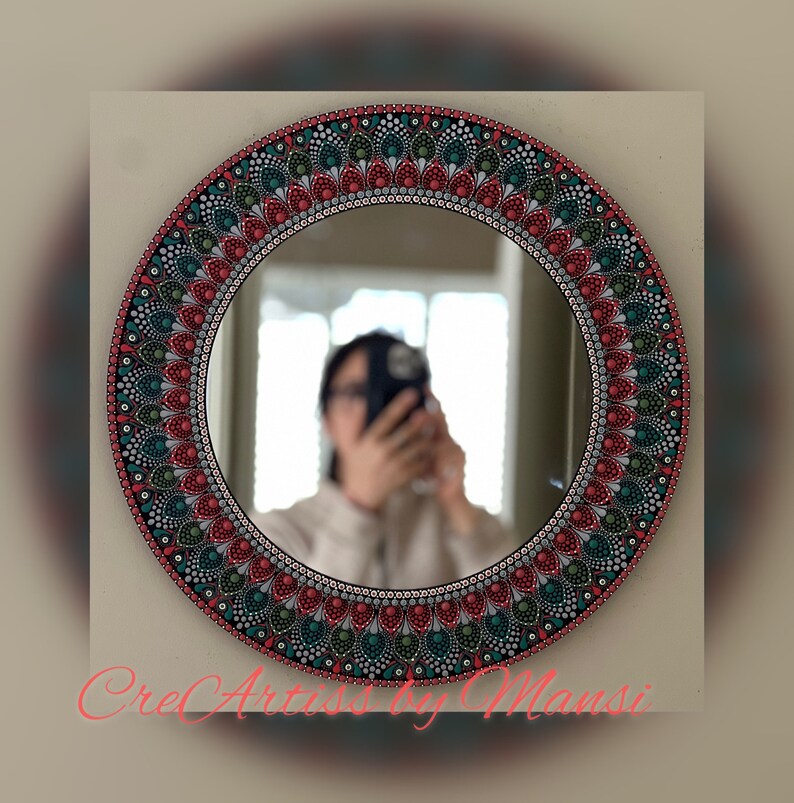 May include: A round mirror with a hand-painted mandala design in shades of red, green, and white. The mirror is surrounded by a black frame.