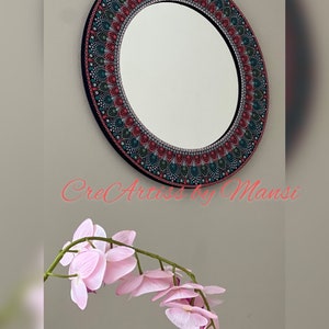 May include: A round mirror with a black frame and a colorful mandala design painted around the edge. The mandala features shades of red, green, and blue. The mirror is hanging on a wall with a pink orchid flower in the foreground.