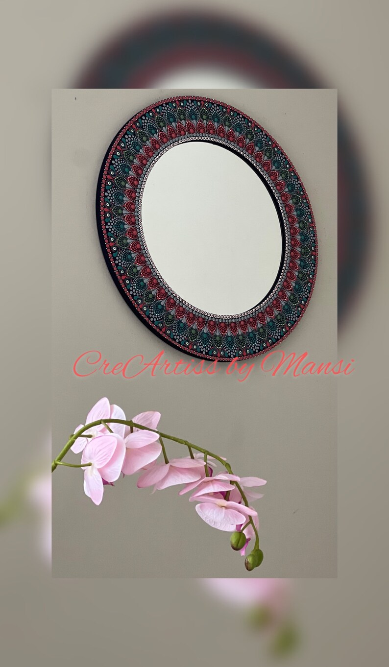 May include: A round mirror with a black frame and a colorful mandala design painted around the edge. The mandala features shades of red, green, and blue. The mirror is hanging on a wall with a pink orchid flower in the foreground.