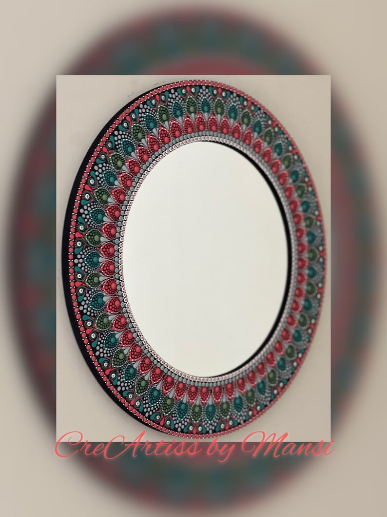 May include: A round mirror with a hand-painted mandala design in shades of red, green, and gray. The mirror is surrounded by a black frame.