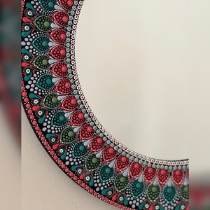 May include: A round mirror with a black frame decorated with a colorful mandala design. The mandala features red, green, and white dots arranged in a circular pattern.