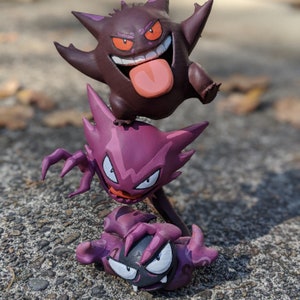 Könnte beinhalten: Drei lila und schwarze Pokémon-Figuren, übereinander gestapelt. Die oberste Figur ist ein Gengar, die mittlere Figur ist ein Hypno und die unterste Figur ist ein Traumato.