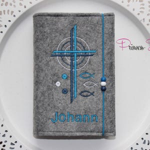 May include: Grey felt book cover with blue and white embroidery of a cross, fish, and the name "Johann".