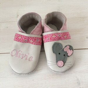 Könnte beinhalten: Ein Paar weiße Lederschuhe für Babys mit rosa Besatz und einem rosa Herzmuster. Ein Schuh hat den Namen "Olivia" bestickt, der andere Schuh hat eine graue Maus-Applikation mit rosa Nase und Schleife.