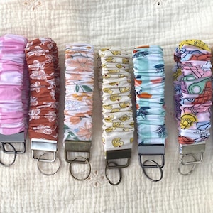 Op de afbeelding: Een verzameling stoffen pols-sleutelhangers in verschillende patronen. Ontwerpen omvatten bloemen-, paisley- en fruitprints. Elke sleutelhanger heeft een metalen ring en een stoffen lus.