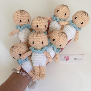 Puede incluir: Un grupo de siete muñecas de crochet que representan bebés, con piel marrón claro, ropa blanca y cuellos con volantes azules. Las muñecas están dispuestas en una mano, con las caras de las muñecas mirando hacia la cámara.