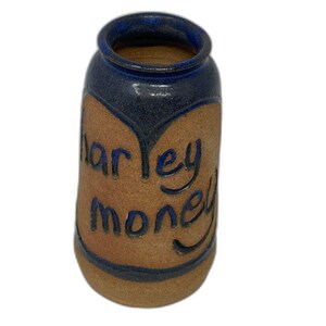 May include: A handmade ceramic jar with a blue rim and base, and the words "Harley Money" in blue script. The jar has a brown, textured surface with a heart-shaped design.