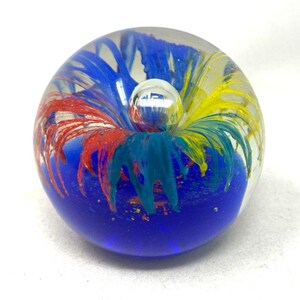May include: A spherical glass paperweight featuring a clear outer layer and an internal design of swirling red, yellow, and blue plumes. The base is a solid deep blue, and a small clear bubble is suspended near the top center of the sphere.