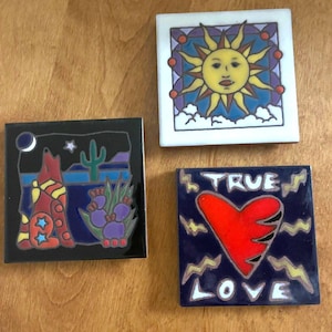 May include: Three square ceramic tiles with colorful designs. One features a sun with a face, another a howling wolf in a desert scene, and the third a red heart with the words "TRUE LOVE".