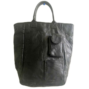 May include: A black tote bag constructed from multiple stitched panels of leather. The bag features two top handles, a large vertical rectangular pocket on the left side, a smaller square pocket on the bottom right, and a flap-covered pocket above it.