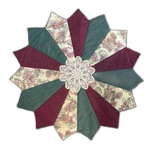 May include: A star-shaped fabric mat featuring alternating panels of solid dark burgundy, solid dark green, and a cream-colored fabric with a printed landscape scene. A circular white crocheted doily is centered on the fabric mat.