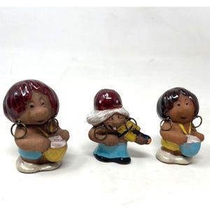 May include: Three ceramic figurines of musicians. Each figure has a brown face, large hoop earrings, and a colorful outfit. One figure plays a violin, while the others play drums. The figurines have red, brown, blue, and yellow accents.