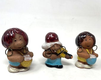 May include: Three ceramic figurines of musicians. Each figure has a brown face, large hoop earrings, and a colorful outfit. One figure plays a violin, while the others play drums. The figurines have red, brown, blue, and yellow accents.