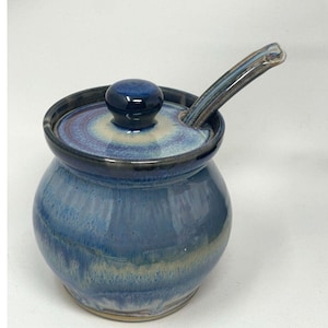 May include: A blue ceramic sugar bowl with a lid and a matching spoon. The bowl has a round shape with a dark blue rim and a lid with a dark blue knob. The spoon is resting on the lid. The bowl has a glazed finish.