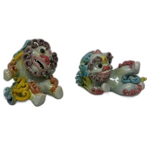 May include: Two ceramic guardian lion statues. One is upright, the other is reclining. Both have detailed features, including curly manes, open mouths, and colorful accents of blue, yellow, and red. The statues are on a white background.