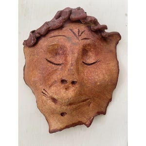 May include: A handmade, ceramic face mask with closed eyes and a serene expression. The mask is primarily brown with darker brown accents for the hair and facial details. The mask has a textured surface and is mounted on a white background.