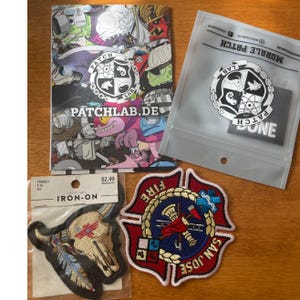 May include: An assortment of iron-on patches and a colorful card. The patches include a bull skull with feathers, a San Jose fire department emblem, and a patch with the text "DONE". The card features cartoon characters and the text "PATCHLAB.DE".