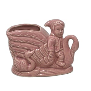 May include: A vintage pink ceramic planter shaped like a swan with a small figure riding on its back. The planter has a large opening for plants and is decorated with detailed swan feathers and a smaller swan on the side.