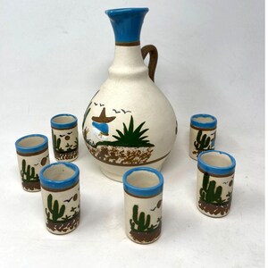 May include: A ceramic liquor set featuring a cream-colored pitcher with a blue top and handle, and six matching shot glasses. The set is decorated with a desert scene with cacti, a person in a sombrero, and birds. The shot glasses have a blue rim.