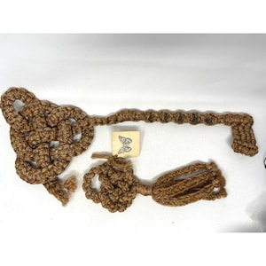 May include: A tan macrame key-shaped wall hanging featuring a large knotted bow head, a long shaft with repeating knots, and a bit at the end. A small square tag with a black butterfly illustration is positioned between the key and a smaller knotted tassel.