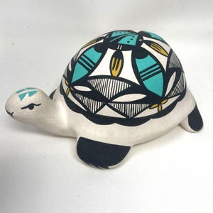 May include: A ceramic turtle figurine with a white body and a patterned shell. The shell features a design of black, turquoise, and yellow geometric shapes. The turtle's head and feet are black and white.