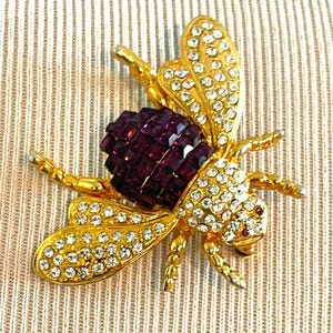 May include: A gold-toned bee brooch with clear crystal accents and a central body of deep purple square-cut stones. The wings and body are detailed with sparkling crystals, creating a glamorous insect-themed accessory.