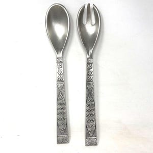 May include: A silver salad serving set, featuring a spoon and a fork. The handles are decorated with floral and geometric patterns. The spoon has an oval bowl, while the fork has three tines. The set is designed for serving salads and other dishes.