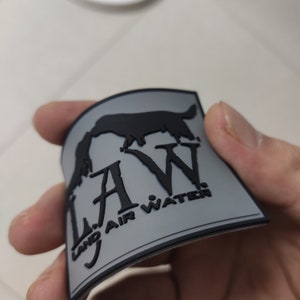 Può includere: Una toppa grigia con una silhouette nera di un cane e il testo "L.A.W. Land Air Water" in nero.
