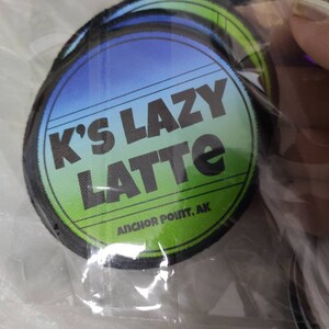 Może przedstawiać: Okrągła haftowana naszywka z tłem przechodzącym od niebieskiego do zielonego. Napis K'S LAZY LATTE jest wydrukowany pogrubionymi czarnymi literami, a poniżej mniejszym czarnym drukiem widnieje ANCHOR POINT, AK. Naszywka ma czarną obszytą krawędź.