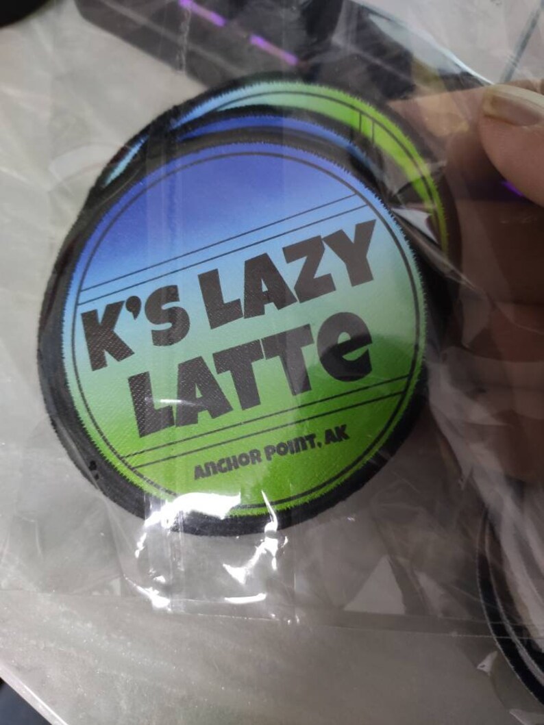 Może przedstawiać: Okrągła haftowana naszywka z tłem przechodzącym od niebieskiego do zielonego. Napis K'S LAZY LATTE jest wydrukowany pogrubionymi czarnymi literami, a poniżej mniejszym czarnym drukiem widnieje ANCHOR POINT, AK. Naszywka ma czarną obszytą krawędź.