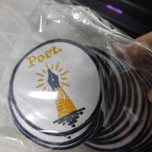 Może przedstawiać: Stos okrągłych haftowanych naszywek w przezroczystej plastikowej torebce. Każda naszywka ma białe tło z ciemnoniebieską obw&oacute;dką, przedstawiającą złotą stal&oacute;wkę pi&oacute;ra z promieniami oraz słowo Poet. wyhaftowane złotą nicią.