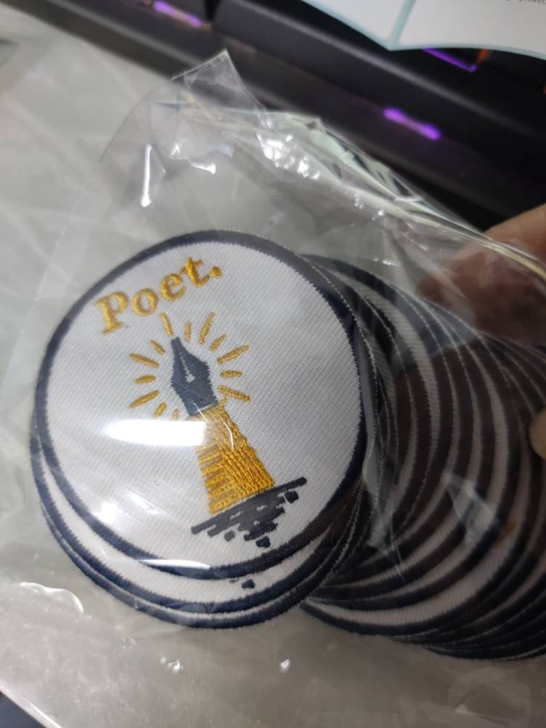 Może przedstawiać: Stos okrągłych haftowanych naszywek w przezroczystej plastikowej torebce. Każda naszywka ma białe tło z ciemnoniebieską obw&oacute;dką, przedstawiającą złotą stal&oacute;wkę pi&oacute;ra z promieniami oraz słowo Poet. wyhaftowane złotą nicią.