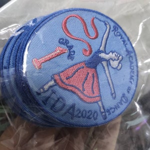 Może przedstawiać: Stos okrągłych haftowanych naszywek w kolorze jasnoniebieskim z ciemnoniebieską obw&oacute;dką. Wz&oacute;r przedstawia tancerza w r&oacute;żowej sp&oacute;dnicy, czerwoną cyfrę 1 oraz wyhaftowany ciemnoniebieski napis Royal Academy of Dance, Grade 1 i IDA 2020.