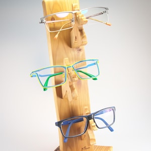 Könnte beinhalten: Ein Brillenständer aus Holz mit drei Brillenpaaren. Die Brillen haben silberne, blaue und grüne Rahmen. Der Ständer ist aus hellem Holz gefertigt und hat ein einzigartiges Schnitzdesign.
